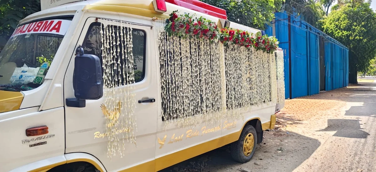 Side profile of Last Ride Funeral Force Traveller premium hearse van with dense jasmine garland side curtains and a crown of red roses along the roof edge — Delhi